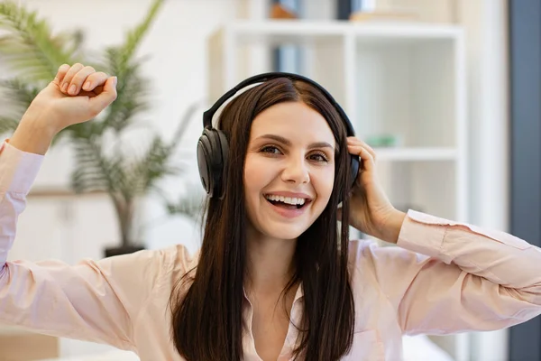 Happy lady in headphones sitting on comfy bed and enjoying relaxing music on weekend. Portrait of happy pretty woman using modern device listening favorite music at home.