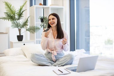 Rahat bir yatakta otururken sesli mesajlaşmaktan hoşlanan gündelik kıyafetli hoş bir kadın. Modern akıllı telefondan konuşmaları kaydederken, evde kalan beyaz kadın hareketi..