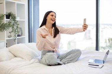 Kablosuz akıllı telefon tutan beyaz bir kadın video görüşmesi yapıyor. Güzel bayan rahat bir yatakta oturuyor ve hafta sonları akrabaları veya arkadaşlarıyla çevrimiçi sohbetin tadını çıkarıyor..