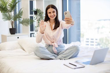 Güzel bayan rahat bir yatakta oturuyor ve hafta sonları akrabaları ya da arkadaşlarıyla çevrimiçi sohbetin tadını çıkarıyor. Kafkasyalı genç bir kadın elinde kablosuz akıllı telefon ile bir video görüşmesi yapıyor..