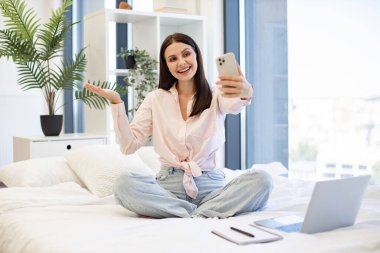 Güzel bayan rahat bir yatakta oturuyor ve hafta sonları akrabaları ya da arkadaşlarıyla çevrimiçi sohbetin tadını çıkarıyor. Kafkasyalı genç bir kadın elinde kablosuz akıllı telefon ile bir video görüşmesi yapıyor..