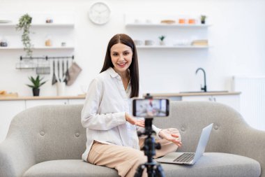 Modern akıllı telefondaki takipçilere günlük kıyafet kayıt videosundaki neşeli içerik yapıcının ön görüntüsü. Bilgisayarlı beyaz kadın mutfaktaki rahat koltukta oturuyor..