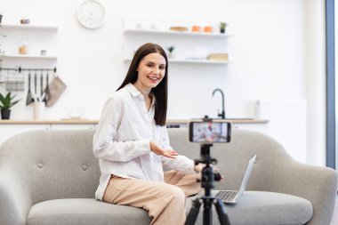 Genç beyaz kadın dizüstü bilgisayarlı mutfaktaki rahat koltukta oturuyor. Modern akıllı telefondaki takipçilere günlük kıyafet kayıt videosundaki neşeli içerik yapıcının ön görüntüsü.