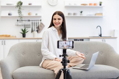 Genç beyaz kadın dizüstü bilgisayarlı mutfaktaki rahat koltukta oturuyor. Modern akıllı telefondaki takipçilere günlük kıyafet kayıt videosundaki neşeli içerik yapıcının ön görüntüsü.