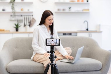 Kafkasyalı kadın etkili video kaydını modern akıllı telefondan dizüstü bilgisayarla koltukta otururken yapıyor. Mutlu genç kadın başarısını internetteki abonelerle paylaşıyor.