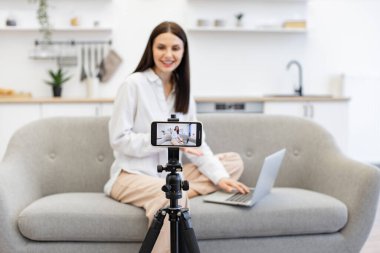 Başarısını internetteki abonelerle paylaşan mutlu genç bir kadın. Modern akıllı telefondaki videoyu kaydeden beyaz kadın etkileyicisi kanepede dizüstü bilgisayarla oturuyor..