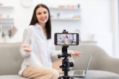 Çevrimiçi eğitim için tripodlu modern akıllı telefon kullanmayı talep ediyor. Beyaz saçlı, esmer bir kadın sosyal ağda canlı yayında modern laptoptan bilgi okuyor..