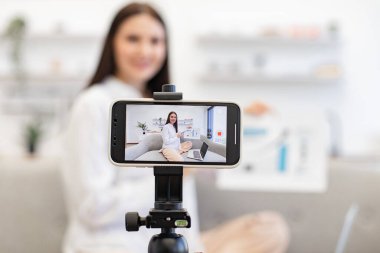 Kafkasyalı esmer finansçı kadın kendini cep telefonuyla kaydediyor. Cep telefonu tripoda tamir ediliyor ve oturma odasındaki abonelere finansal istatistikler gösteriyor. Teknoloji ve blog kavramı.