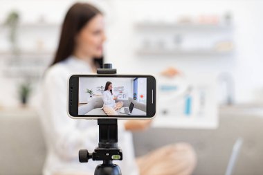 Kafkasyalı esmer finansçı kadın kendini cep telefonuyla kaydediyor. Cep telefonu tripoda tamir ediliyor ve oturma odasındaki abonelere finansal istatistikler gösteriyor. Teknoloji ve blog kavramı.