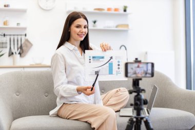 Teknoloji ve blog kavramı. Kafkasyalı esmer finansçı kadın kendini cep telefonuyla kaydediyor. Cep telefonu tripoda tamir ediliyor ve oturma odasındaki abonelere finansal istatistikler gösteriyor..