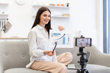 Teknoloji ve blog kavramı. Kafkasyalı esmer finansçı kadın kendini cep telefonuyla kaydediyor. Cep telefonu tripoda tamir ediliyor ve oturma odasındaki abonelere finansal istatistikler gösteriyor..