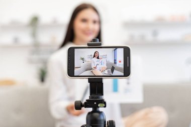 Teknoloji ve blog kavramı. Kafkasyalı esmer finansçı kadın kendini cep telefonuyla kaydediyor. Cep telefonu tripoda tamir ediliyor ve oturma odasındaki abonelere finansal istatistikler gösteriyor..