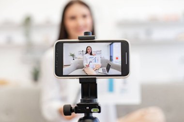 Teknoloji ve blog kavramı. Kafkasyalı esmer finansçı kadın kendini cep telefonuyla kaydediyor. Cep telefonu tripoda tamir ediliyor ve oturma odasındaki abonelere finansal istatistikler gösteriyor..