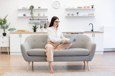 Rahat bir kanepede otururken kablosuz bilgisayarda yazan günlük kıyafetlerle odaklanmış beyaz bir kadın. Çekici esmer kadın serbest yazar evde otururken online iş yapıyor..