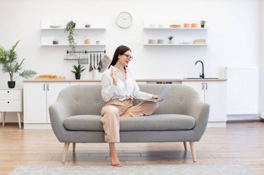 Webcam 'de konuşan ve elleriyle hareket eden kendine güvenen esmer kadın. Kanepede oturan ve evdeki video konferansı için modern dizüstü bilgisayar kullanan çekici kadın serbest yazar..