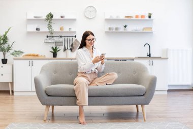 Evden uzakta çalışan mutlu genç bir kadın modern kablosuz cep telefonuyla daktilo çekiyor. Rahat bayan rahat koltukta oturuyor ve hafta sonları akıllı telefon kullanarak iş arkadaşlarıyla sohbet ediyor..