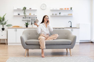 Güzel bir kadın evdeki kanepede dinleniyor ve modern akıllı telefonu ailesiyle konuşmak için kullanıyor. Modern cihazlar kullanan Kafkas esmeri, hafta sonu iş görüşmelerinde mola veriyor..