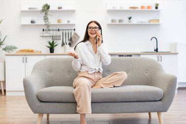 Güzel bir kadın evdeki kanepede dinleniyor ve modern akıllı telefonu ailesiyle konuşmak için kullanıyor. Modern cihazlar kullanan Kafkas esmeri, hafta sonu iş görüşmelerinde mola veriyor..