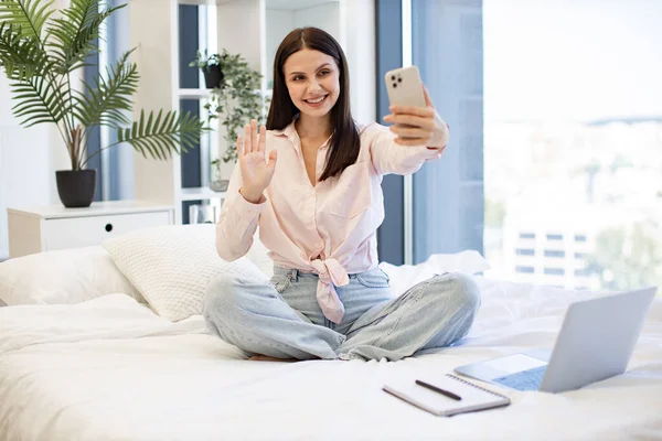 Güzel bayan rahat bir yatakta oturuyor ve hafta sonları akrabaları ya da arkadaşlarıyla çevrimiçi sohbetin tadını çıkarıyor. Kafkasyalı genç bir kadın elinde kablosuz akıllı telefon ile bir video görüşmesi yapıyor..