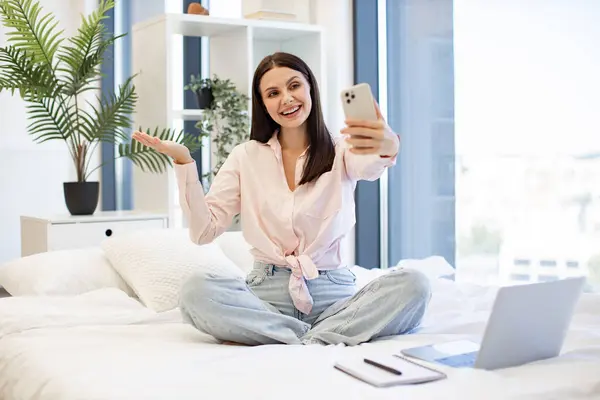 Güzel bayan rahat bir yatakta oturuyor ve hafta sonları akrabaları ya da arkadaşlarıyla çevrimiçi sohbetin tadını çıkarıyor. Kafkasyalı genç bir kadın elinde kablosuz akıllı telefon ile bir video görüşmesi yapıyor..