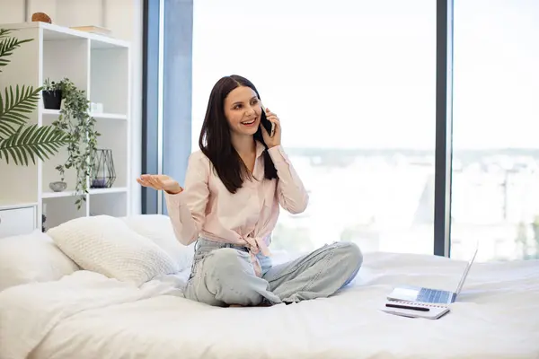 Beautiful woman relaxing on bed at home and using modern smartphone for talking with family. Caucasian brunette using modern devices enjoying break from work for business negotiations on weekend.
