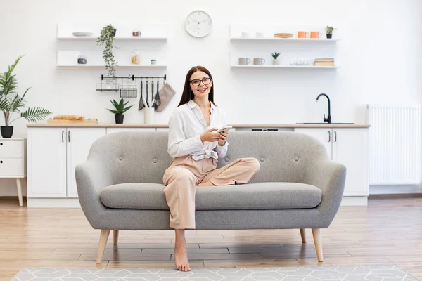 Relaxed lady sitting on comfy sofa and enjoying chatting with coworkers using smartphone on weekend. Happy young woman working remotely from home, typing on modern wireless cell phone.