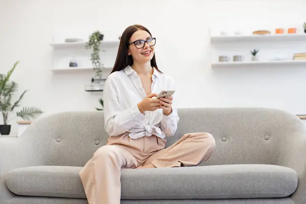 Charming woman in casual attire enjoying exchanging of sms messages while sitting on cozy couch. Positive caucasian female typing text for her conversation on modern smartphone, staying at home.