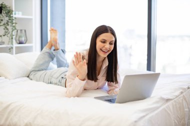 Modern dizüstü bilgisayarla konuşan ve evde kalan esmer bayanın selamlaşması. Rahat bir yatakta uzanırken ailemle çevrimiçi sohbetin tadını çıkaran gündelik kıyafetli hoş bir kadın..