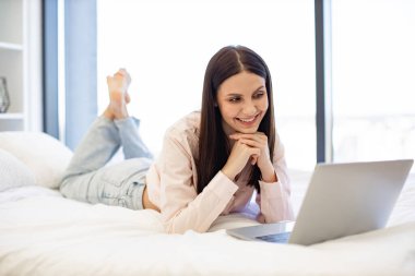 Mutlu gülümseyen esmer kadın, modern dizüstü bilgisayar kullanarak film izliyor, evde kalıyor. Rahat bir yatakta uzanırken internette bilgi okumaktan zevk alan gündelik kıyafetli hoş bir kadın..