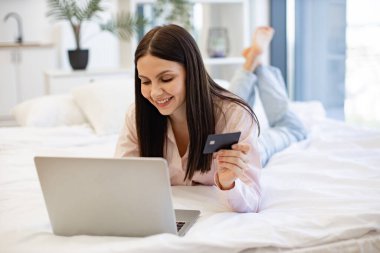 Modern dizüstü bilgisayarda yazan çekici esmer kadın kredi kartı bilgilerini satın almak için giriş yapıyor, evde kalıyor. Rahat bir yatakta uzanırken online alışverişin tadını çıkaran gündelik kıyafetli hoş bir kadın..