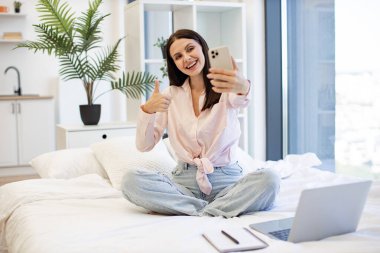 Kafkasyalı genç bir kadın elinde kablosuz akıllı telefon ile baş parmağını gösteren bir video görüşmesi yapıyor. Güzel bayan rahat bir yatakta oturuyor ve hafta sonları akrabaları veya arkadaşlarıyla çevrimiçi sohbetin tadını çıkarıyor..