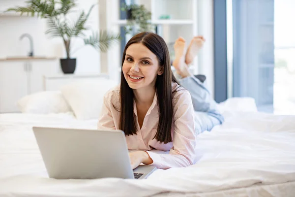 Charming woman in casual attire enjoying online conversation with family while lying on cozy bed, looking at camera. Brunette female typing message in social media using modern laptop, staying at home