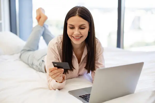 Smiling young woman making online order using modern wireless laptop. Relaxed lady lying on comfy bed and enjoying purchase of goods on Internet using bank card at home on weekend.