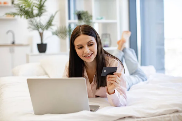 Relaxed lady lying on comfy bed and enjoying purchase of goods on Internet using bank card at home on weekend. Smiling young woman making online order using modern wireless laptop.