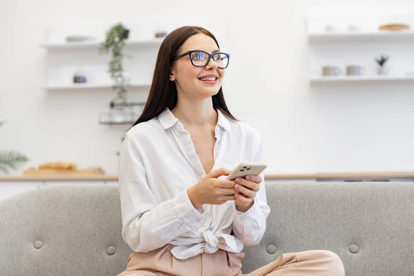 Charming woman in casual attire enjoying exchanging of sms messages while sitting on cozy couch. Positive caucasian female typing text for her conversation on modern smartphone, staying at home.