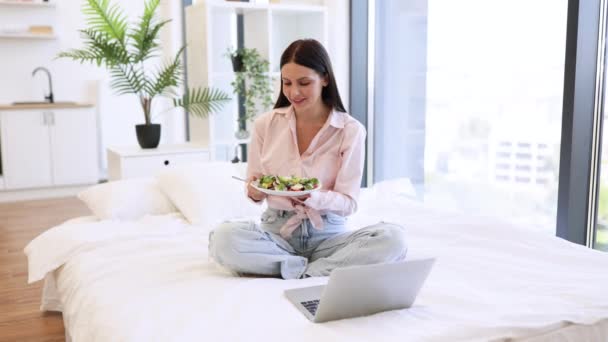 漂亮的年轻女子在舒适的床上悠闲自在地呆在家里 一边用现代笔记本电脑一边吃着健康的沙拉 白种人在周末享用新鲜蔬菜的早餐 — 图库视频影像