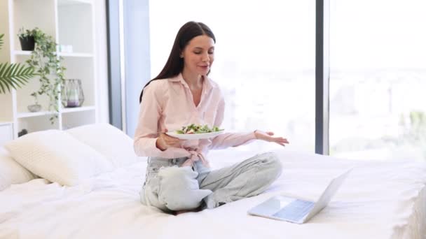 快乐的黑发女性通过视频通话使用笔记本电脑和吃健康的沙拉 迷人的女人穿着休闲装 坐在舒适的床上 享受新鲜蔬菜早餐 待在家里 — 图库视频影像