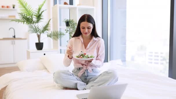年轻女性在舒适舒适的家里放松的形象 在吃健康沙拉的同时使用现代笔记本电脑 白种人的黑头发 周末享受新鲜蔬菜的早餐 看着相机 — 图库视频影像