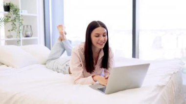 Modern dizüstü bilgisayarla konuşan ve evde kalan esmer bayanın selamlaşması. Rahat bir yatakta uzanırken ailemle çevrimiçi sohbetin tadını çıkaran gündelik kıyafetli hoş bir kadın..
