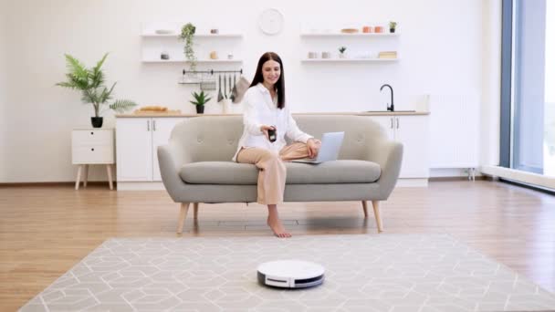 若い白人ブルネット女性ラップトッププログラミングの部屋の床の自動整頓 キッチンでソファーに座っている間 リモートコントロールでロボット掃除機を切り替える美しい女性 — ストック動画