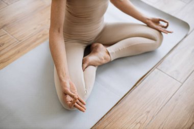 Fiziksel faydalarıyla övünen ileri düzey bir poz. Bej bluzlu ince kadın ve yoga pantolonlu lotus pozisyonu alan parlak spor salonunun arka planında Mudra jesti yapan kadın..