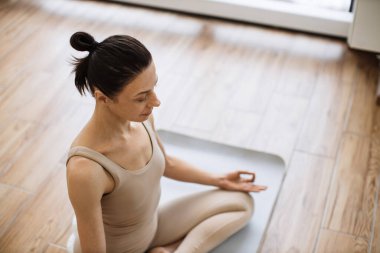 Fiziksel faydalarıyla övünen ileri düzey bir poz. Bej bluzlu ince kadın ve yoga pantolonlu lotus pozisyonu alan parlak spor salonunun arka planında Mudra jesti yapan kadın..