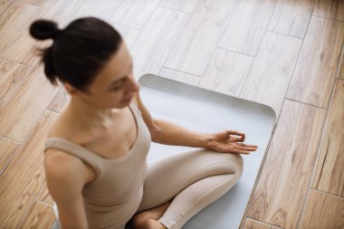 Bej bluzlu ince bir kadın ve parlak spor salonunun arka planında Mudra jesti yaparak nilüfer çiçeği pozisyonuna ulaşan yoga pantolonu. Fiziksel faydalarıyla övünen ileri düzey poz.