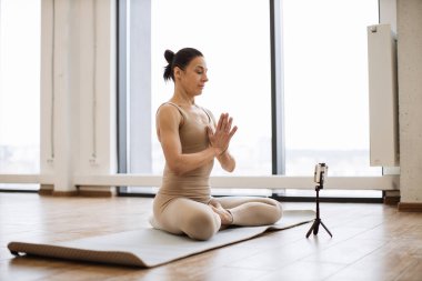 Ünlü blogcu, kendi takipçileri için akıllı telefondan stüdyoda meditasyon hakkında video içeriği çekiyor. Huzurlu Kafkasyalı kadın lotus pozunda asma sana performansı sergiliyor. Sağlıklı bir yaşam tarzına sahip..