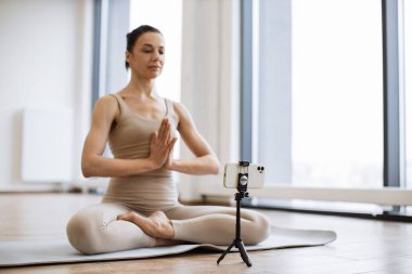 Sportif yetişkinler yoga dersi sırasında akıllı telefondan video çekerek minder üzerinde asma sana egzersizi yapıyorlar. Son sınıf bayan blog yazarı spor kıyafetleriyle stüdyoda bacak bacak bacak üstüne atarak meditasyon yapıyor..