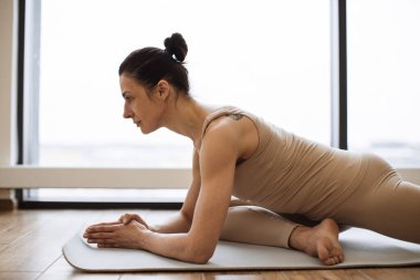 Asya 'da yoga minderinde uzanan bej rengi spor giysili Kafkasyalı esmer, arka planda parlak fitness merkezinde panoramik pencereler..