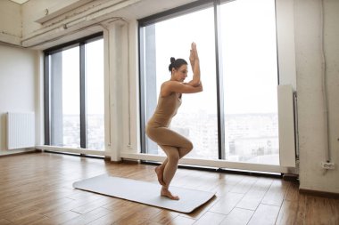 Spor kıyafetleri içinde beyaz bir kadın arka planda Fransız pencereleriyle yoga pozu veriyor. Güçlü, aktif bir kadın tek bacağıyla dengede duruyor ve antrenman odasında el ele tutuşuyor..