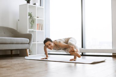 Güçlü, çekici bir kadın elleri üzerinde duruyor ve içeride yoga yaparken bacaklarını yukarıda tutuyor. Spor kıyafetli aktif esmer çeşitli pozisyonlarda vücut dayanıklılığı ve denge egzersizleri yapıyor..