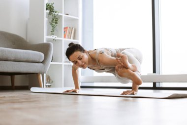 Güçlü, çekici bir kadın elleri üzerinde duruyor ve içeride yoga yaparken bacaklarını yukarıda tutuyor. Spor kıyafetli aktif esmer çeşitli pozisyonlarda vücut dayanıklılığı ve denge egzersizleri yapıyor..
