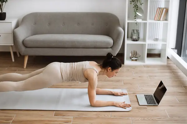 Mature female strengthening whole body with plank pose during yoga workout practice during onine class using laptop at home. Sporty woman in activewear improving posture with Phalakasana II exercise.
